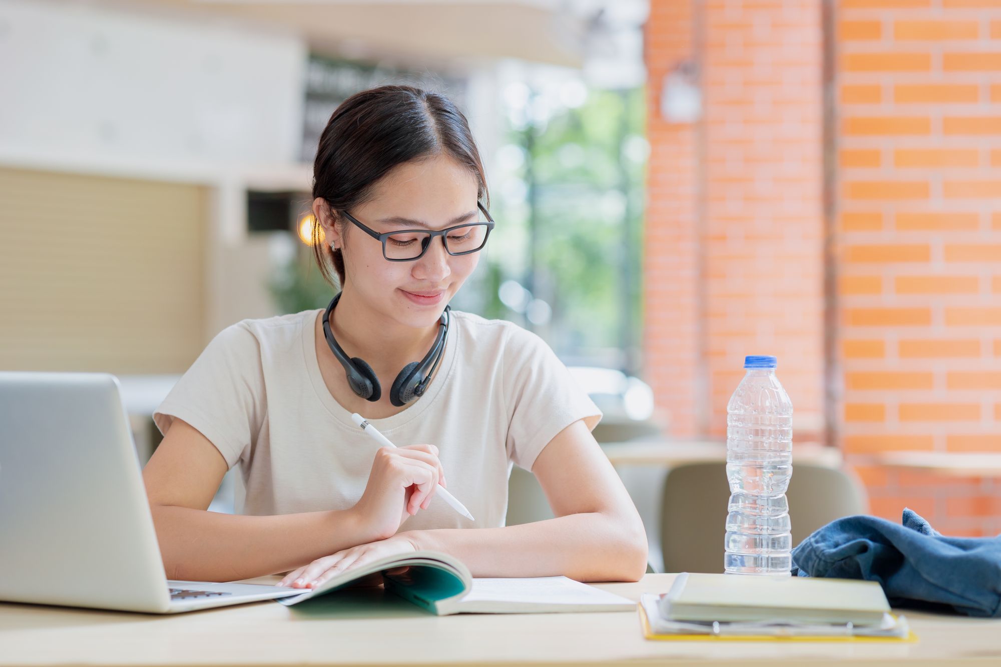 Young woman with glasses reading and taking notes at a desk with a laptop and notebook