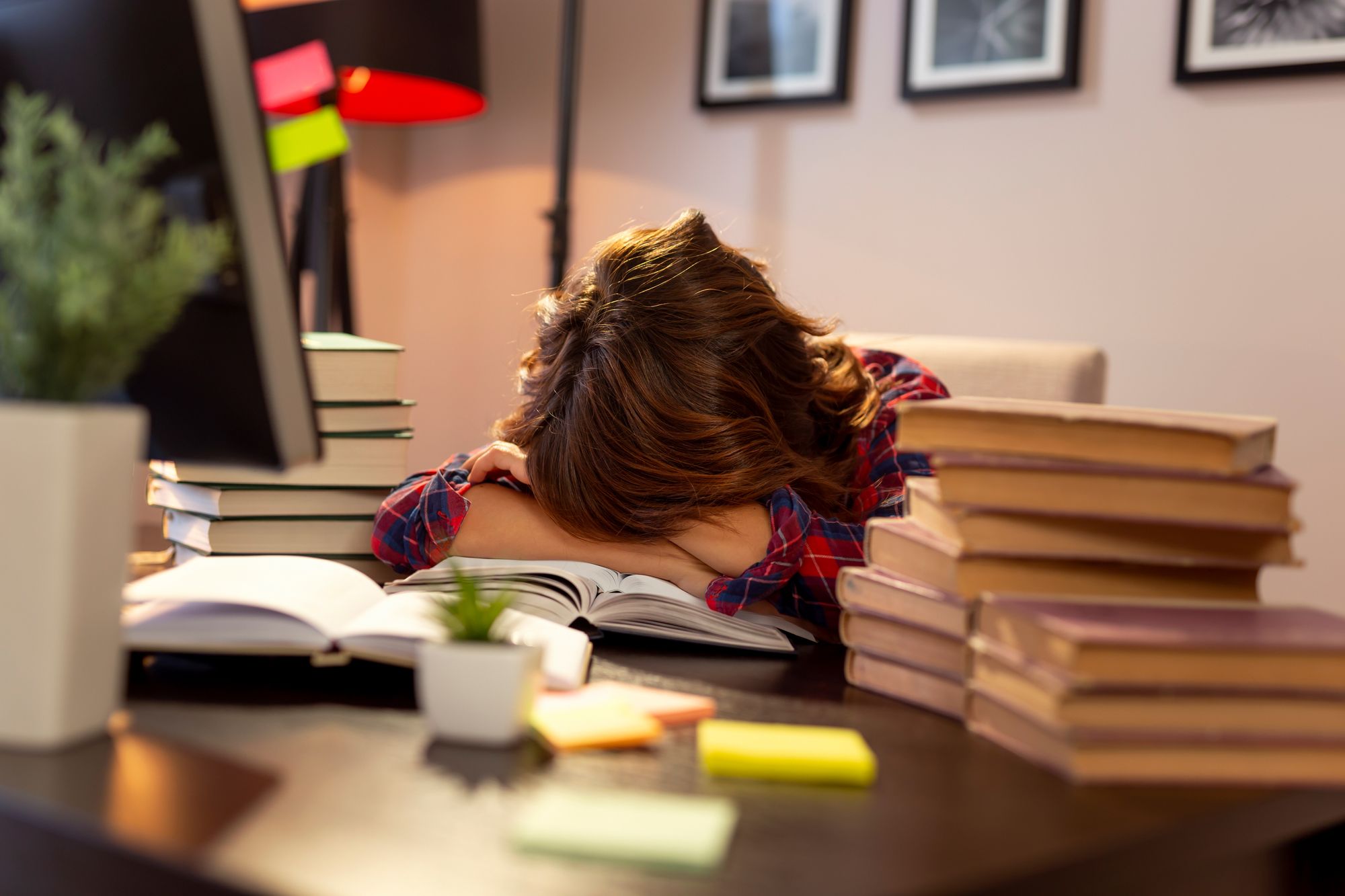 Exhausted female student asleep on an open book surrounded by stacks of study materials