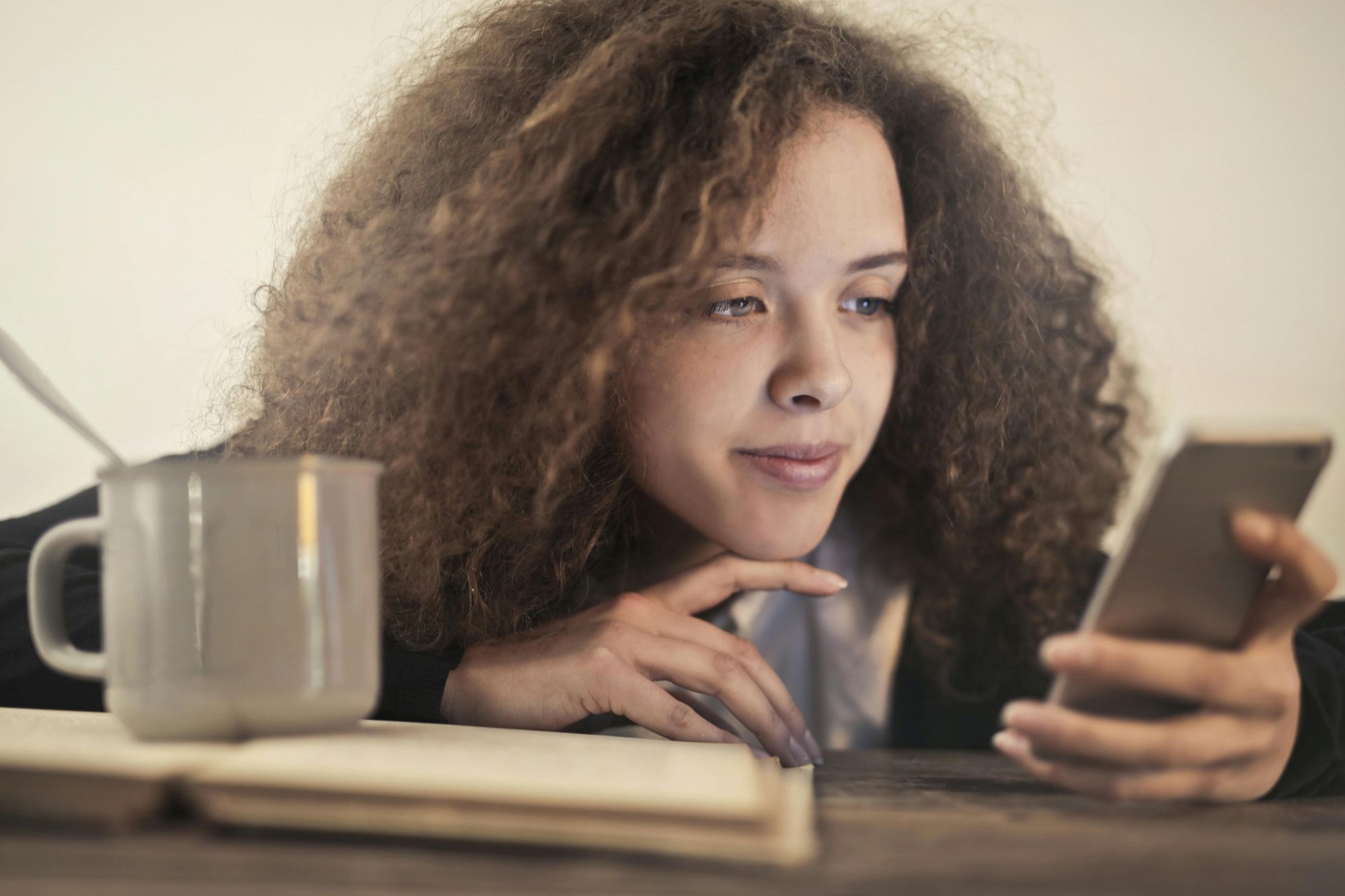 Woman checking her phone with a cup of coffee and an open book in front of her