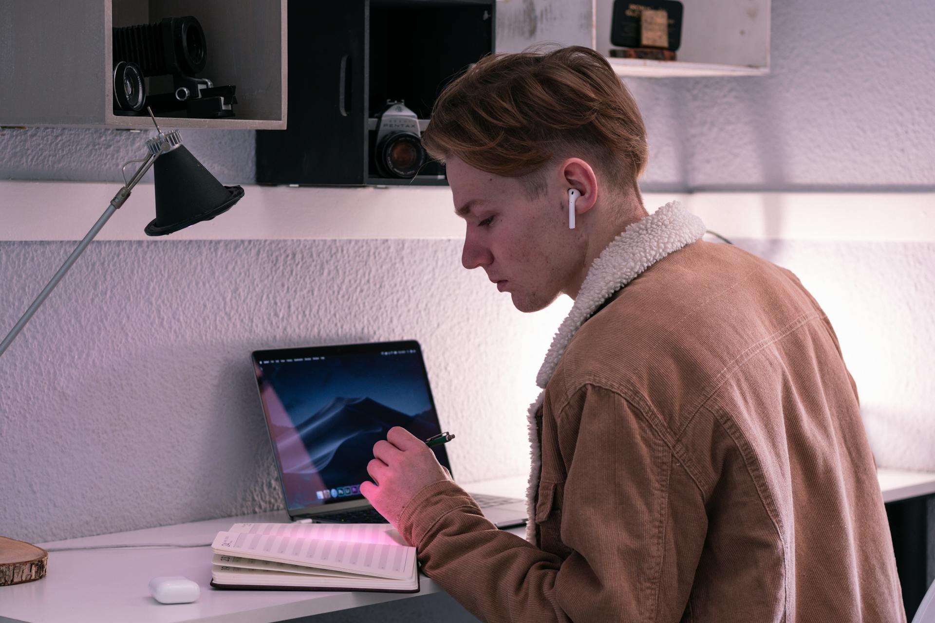 A person sitting at the desk with a pen in hand, and a notebook and laptop on the desk