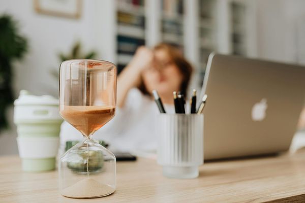 Hourglass in the foreground as a woman pauses from working on a laptop in the background